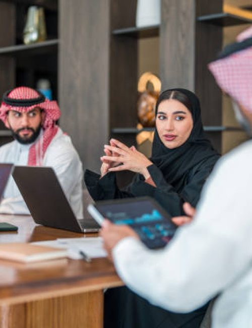 Diverse Arabic business executives discuss strategies at a meeting. The man wears traditional attire, while the woman is in a black abaya. Laptops and documents are present in a modern office setting, highlighting professional collaboration and analysis.